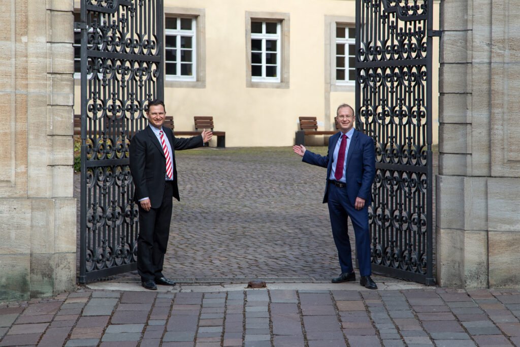 Certified Financial Modeler 5 Zwei Professoren in Anzügen stehen an einem geöffneten schmiedeeisernen Tor und laden symbolisch in den Innenhof eines historischen Gebäudes ein.
