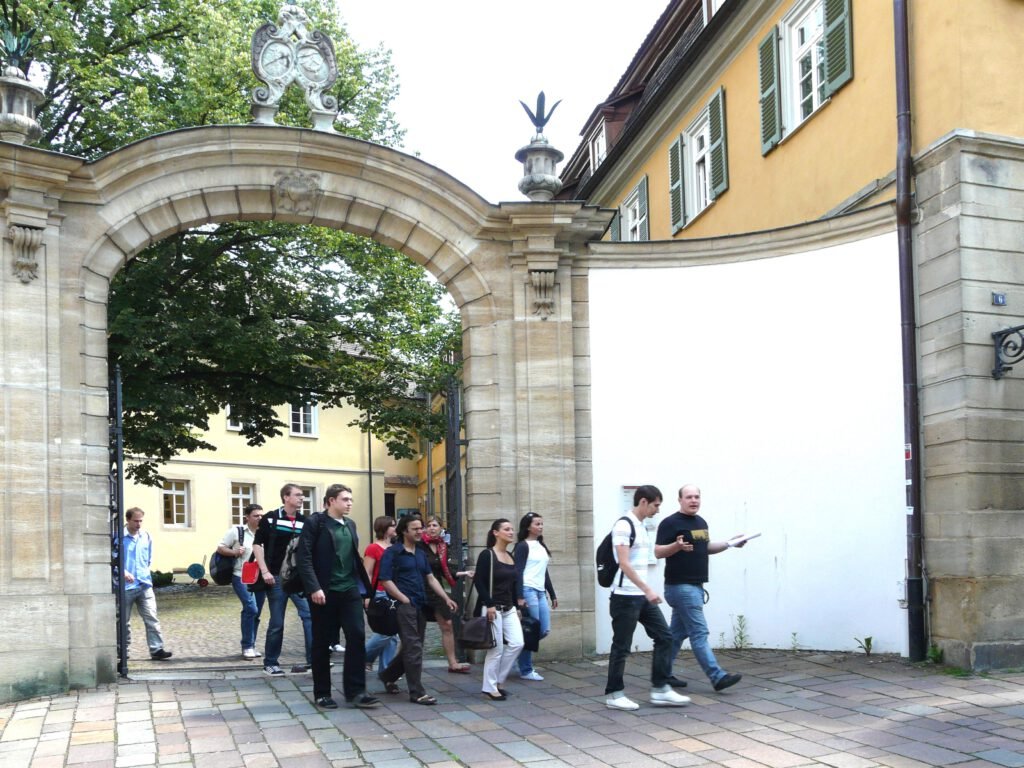 Certified Financial Modeler 3 Eine Gruppe von Studierenden betritt durch ein historisches Tor einen Innenhof mit barocker Architektur. Ein Mann mit Notizbuch führt die Gruppe an.